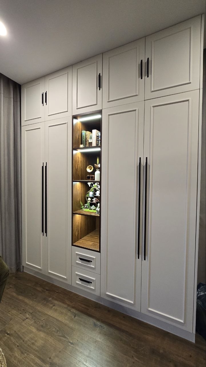Real bedroom wardrobe with white panel doors, dark handles, wood-toned display shelves, lighting and drawers